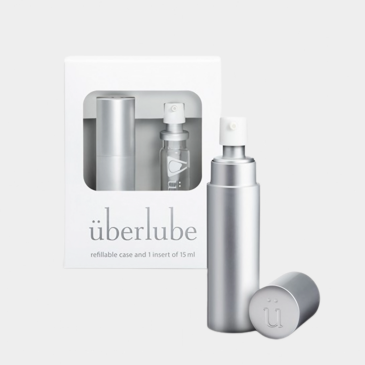 A silver-colored personal lubricant dispenser with a removable cap and a 15ml lube insert displayed alongside its packaging box which reads 'ubælube'. The product is described as refillable and designed for travel. — 1