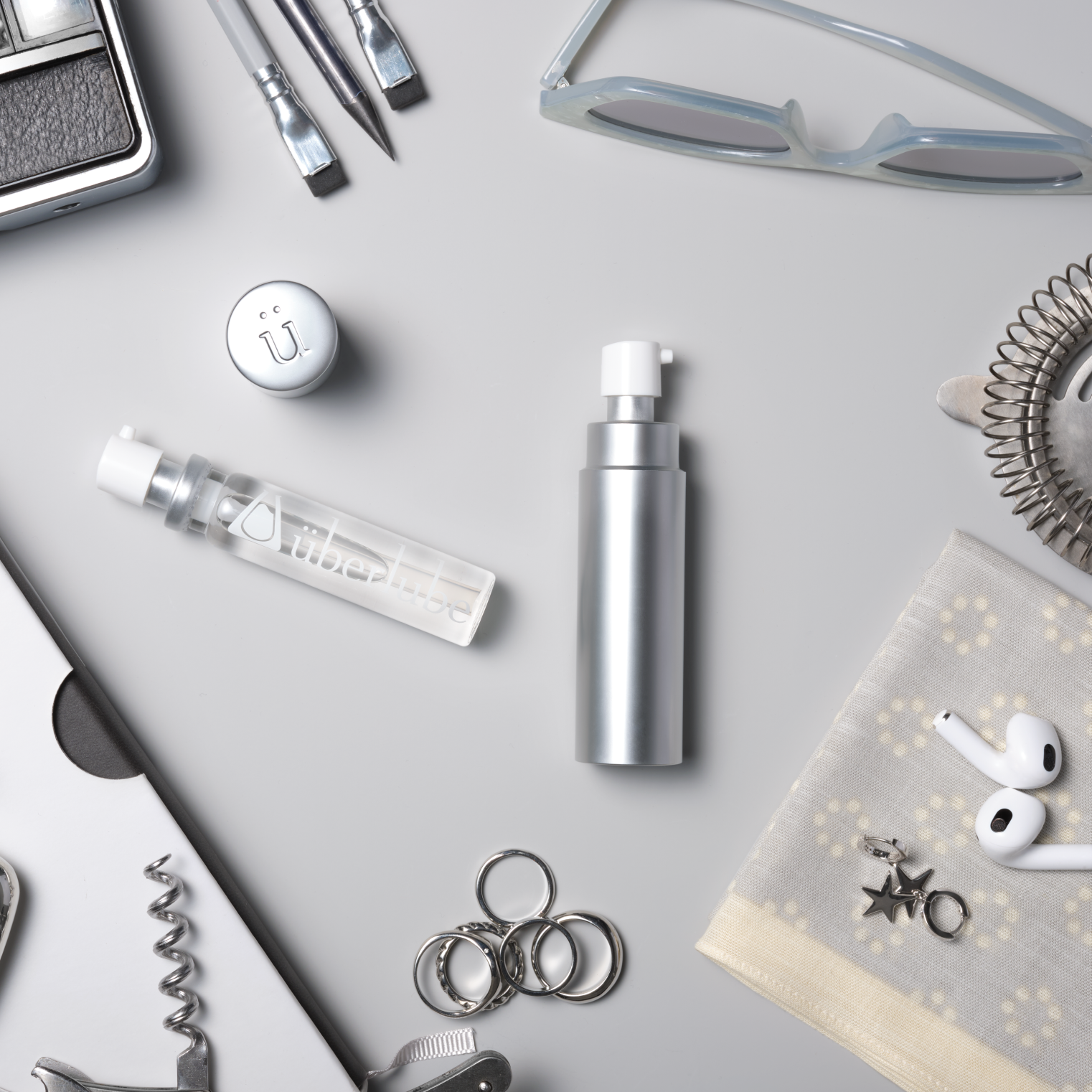 Collection of silver items around an uberlube silver travel including a 15ml vial, pen, and glasses on a light gray surface.